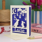 A laser cut 1st birthday card with cut out bunnies looking up at the moon and stars, personalised with the age 1 in a cloud in the sky, and the name "Lacie" at the bottom. 