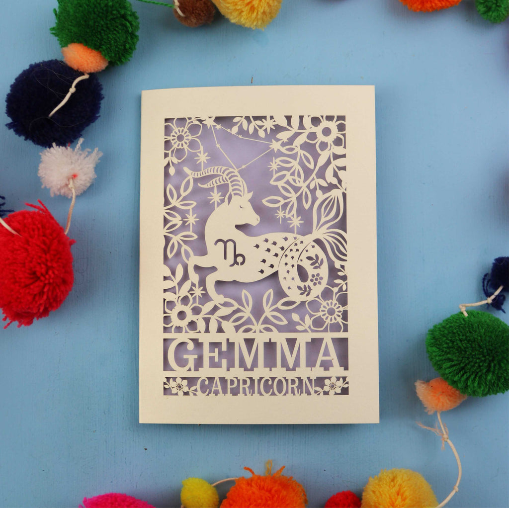 Decorative birthday card with Capricorn zodiac symbol on a blue background with colourful pom-poms.