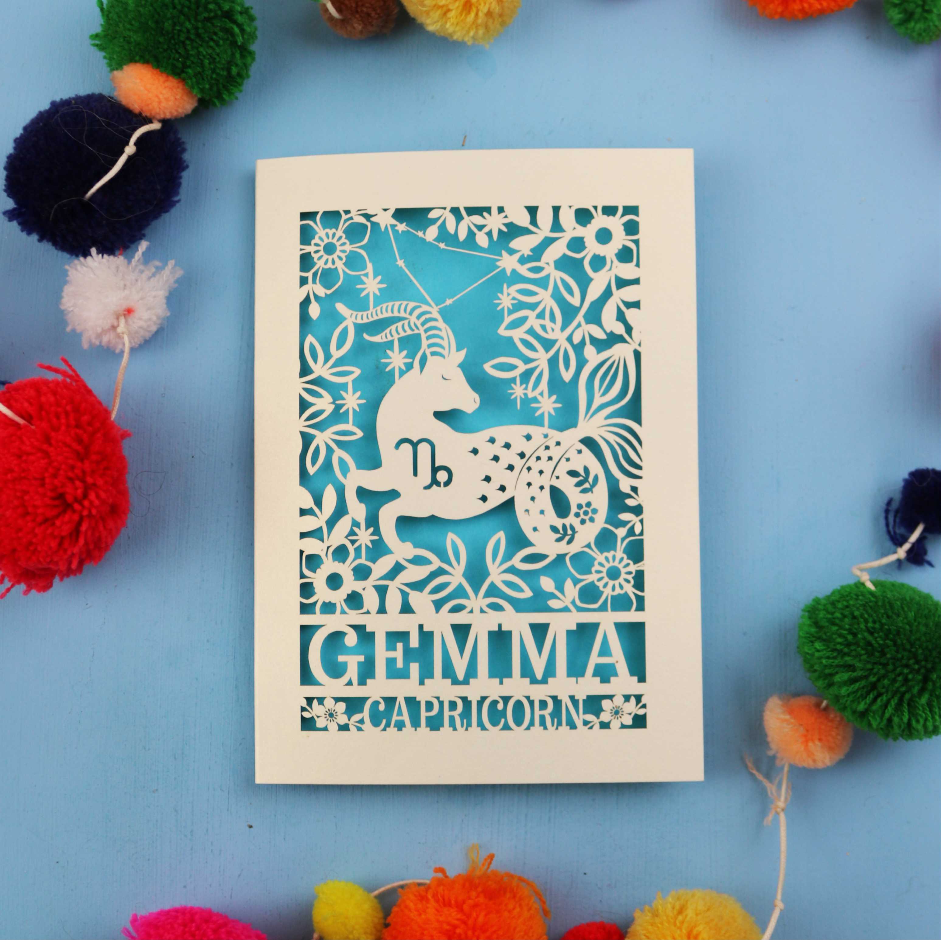 Decorative birthday card with Capricorn zodiac symbol on a blue background with colourful pom-poms.