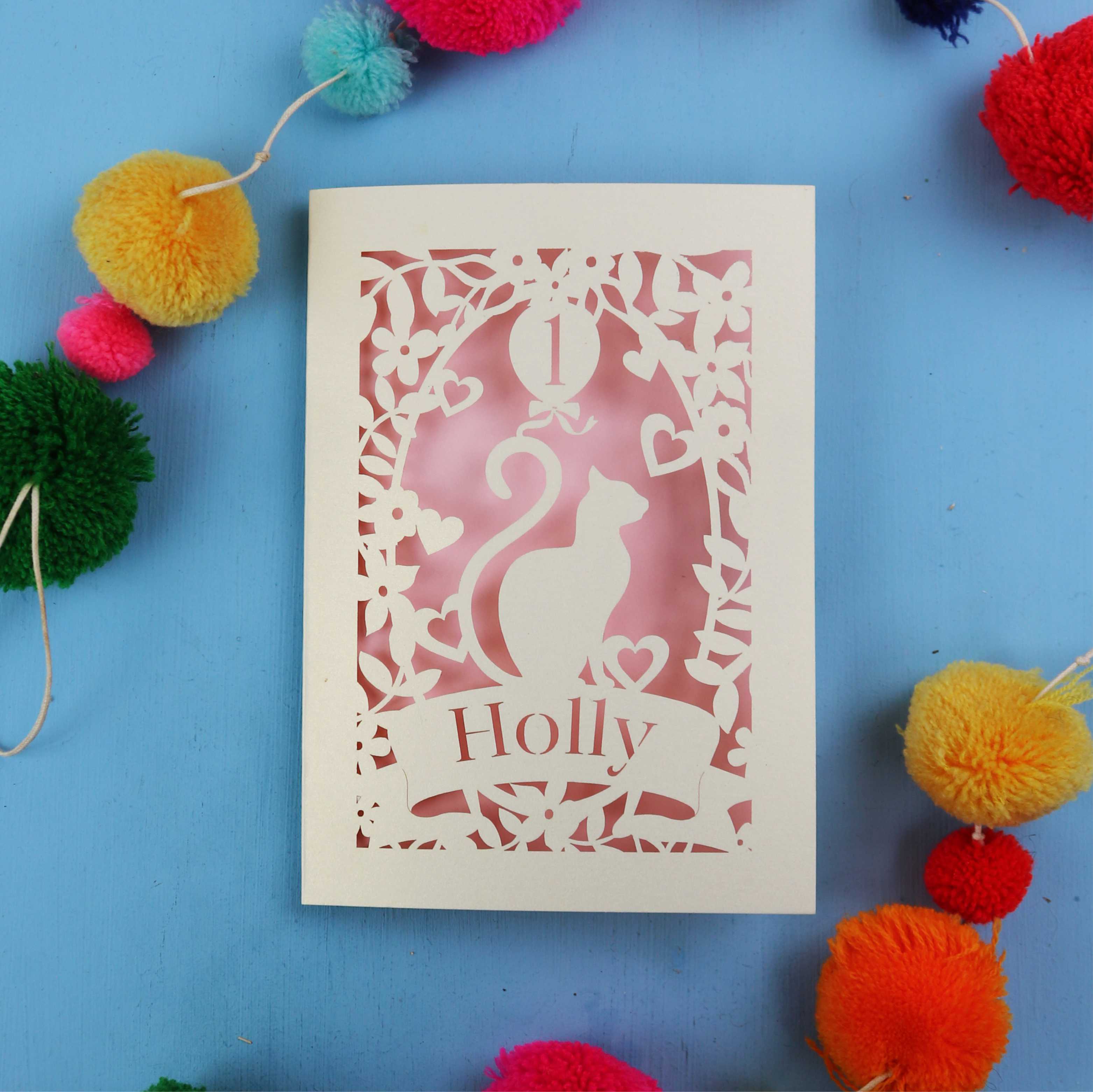 First birthday card with cut-out design of a cat and 'Holly' text, surrounded by colourful pom-poms on a blue background.