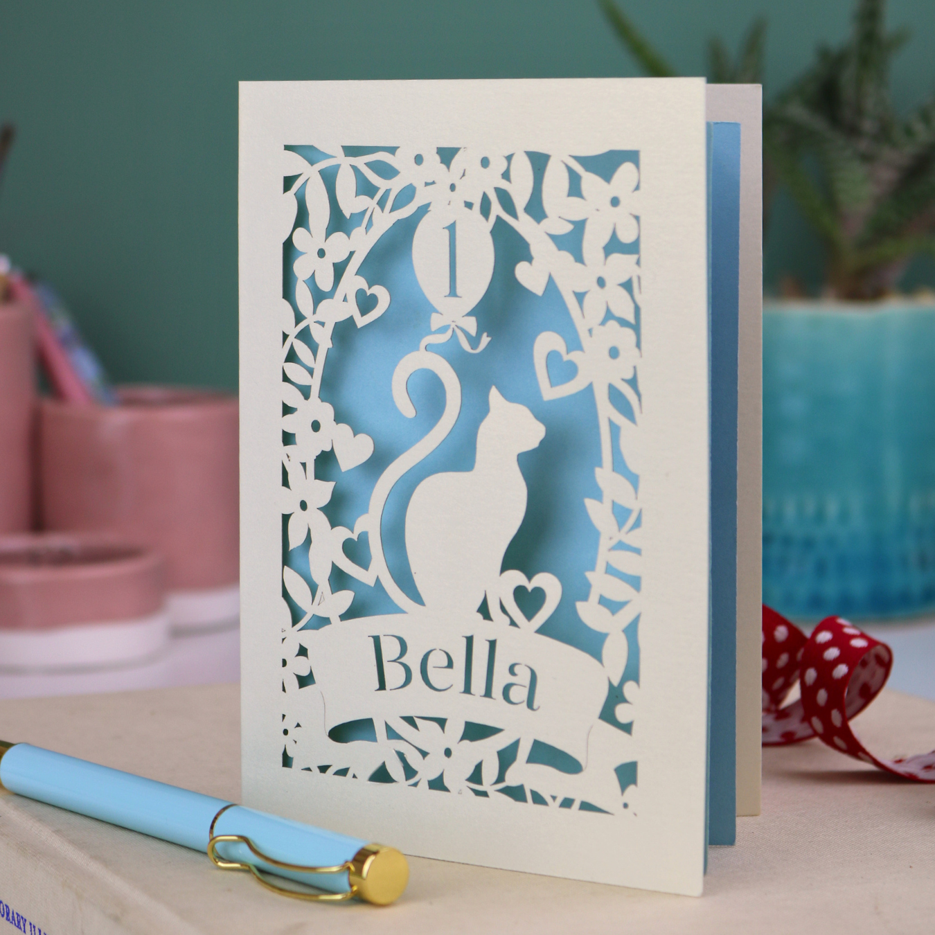 Laser cut first birthday card with a cat silhouette and 'Bella' text, placed on a surface with decorative items.