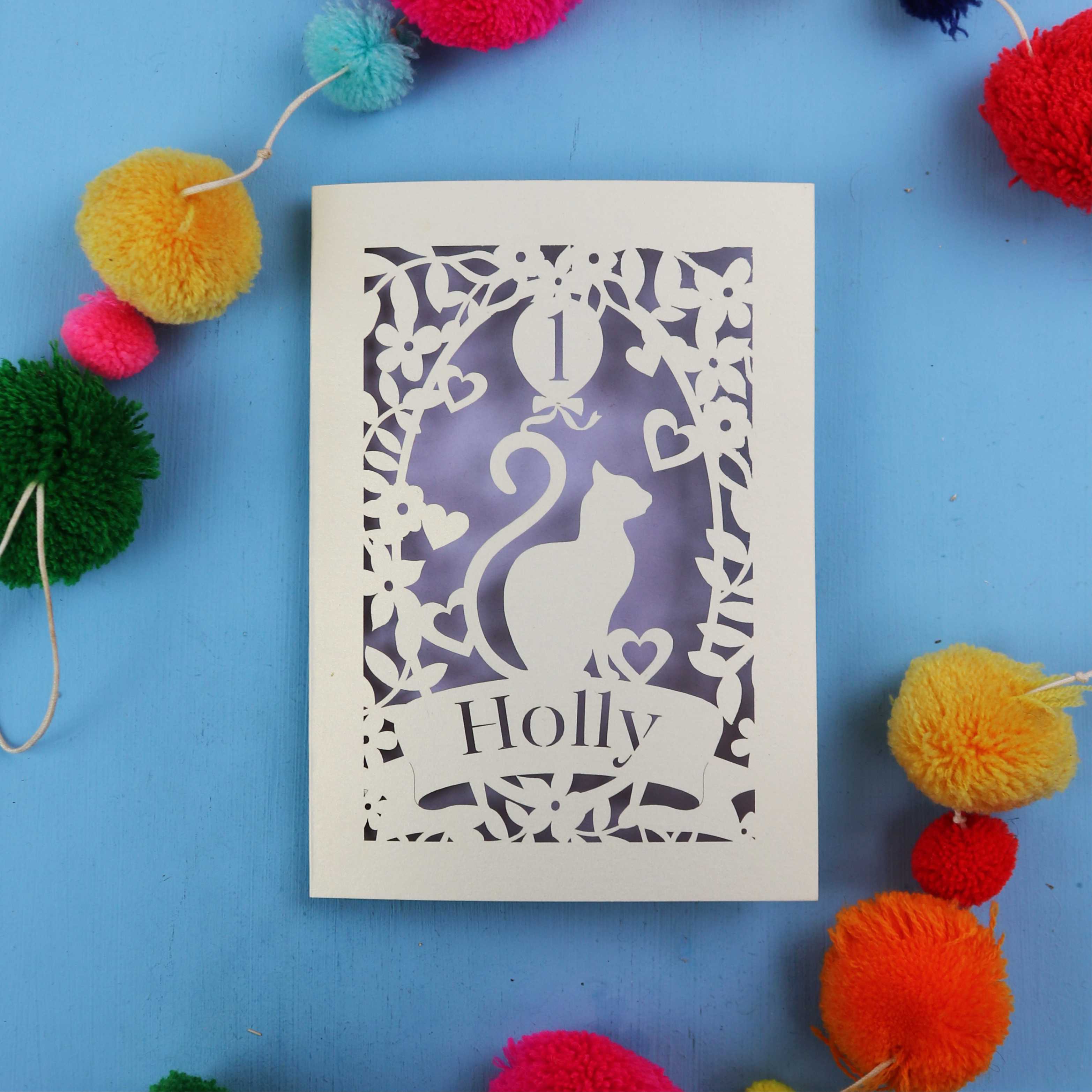 Decorative 1st birthday card with cut-out design of a cat and 'Holly' text, surrounded by colourful pom-poms on a blue background.