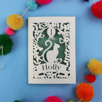 Decorative 1st birthday card with cut-out design of a cat and 'Holly' text, surrounded by colourful pom-poms on a blue background.
