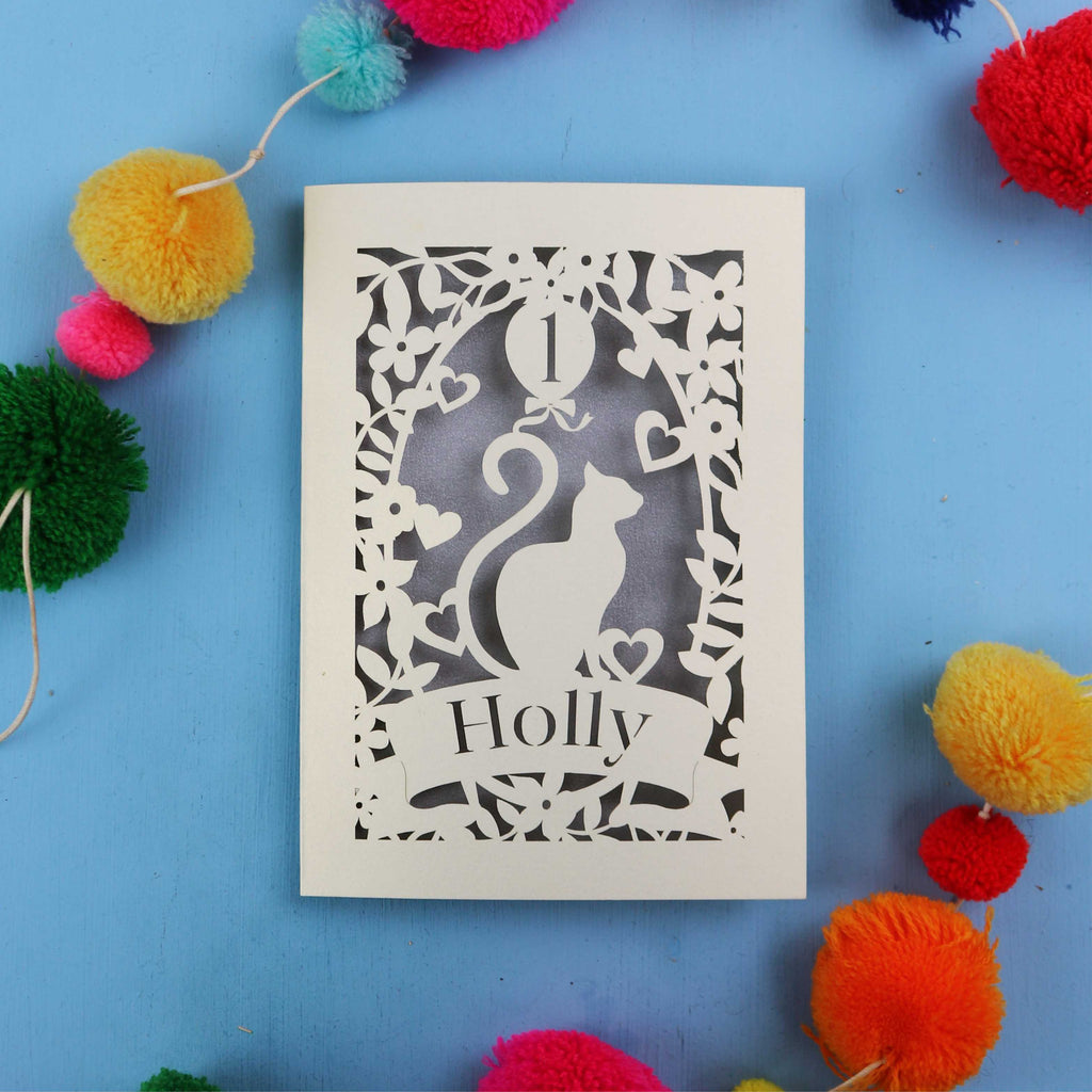 Decorative Laser cut 1st birthday card with cut-out design of a cat and 'Holly' text, surrounded by colourful pom-poms on a blue background.