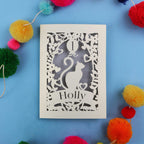 Decorative Laser cut 1st birthday card with cut-out design of a cat and 'Holly' text, surrounded by colourful pom-poms on a blue background.