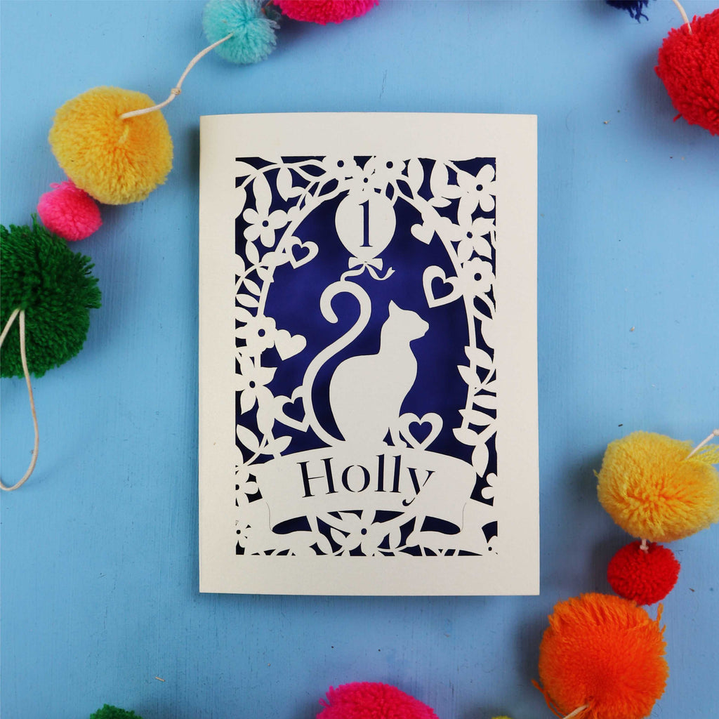 Decorative 1st birthday card with cut-out design of a cat and 'Holly' text, surrounded by colourful pom-poms on a blue background.