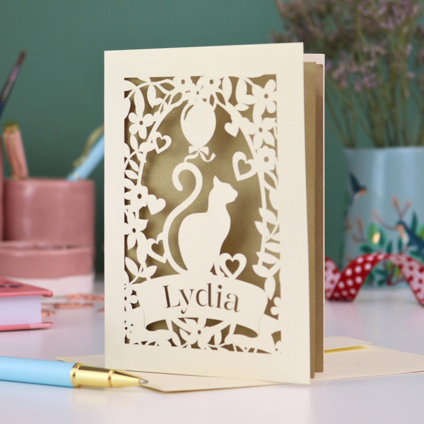 A laser cut birthday card with cat design and personalised with a name, on a desk with stationery items. 