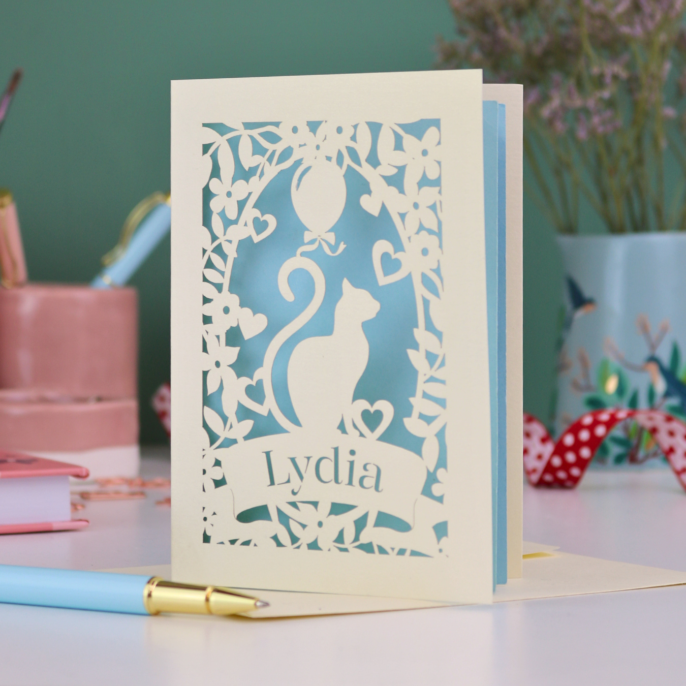 A laser cut birthday card with cat design and personalised with a name, on a desk with stationery items. 
