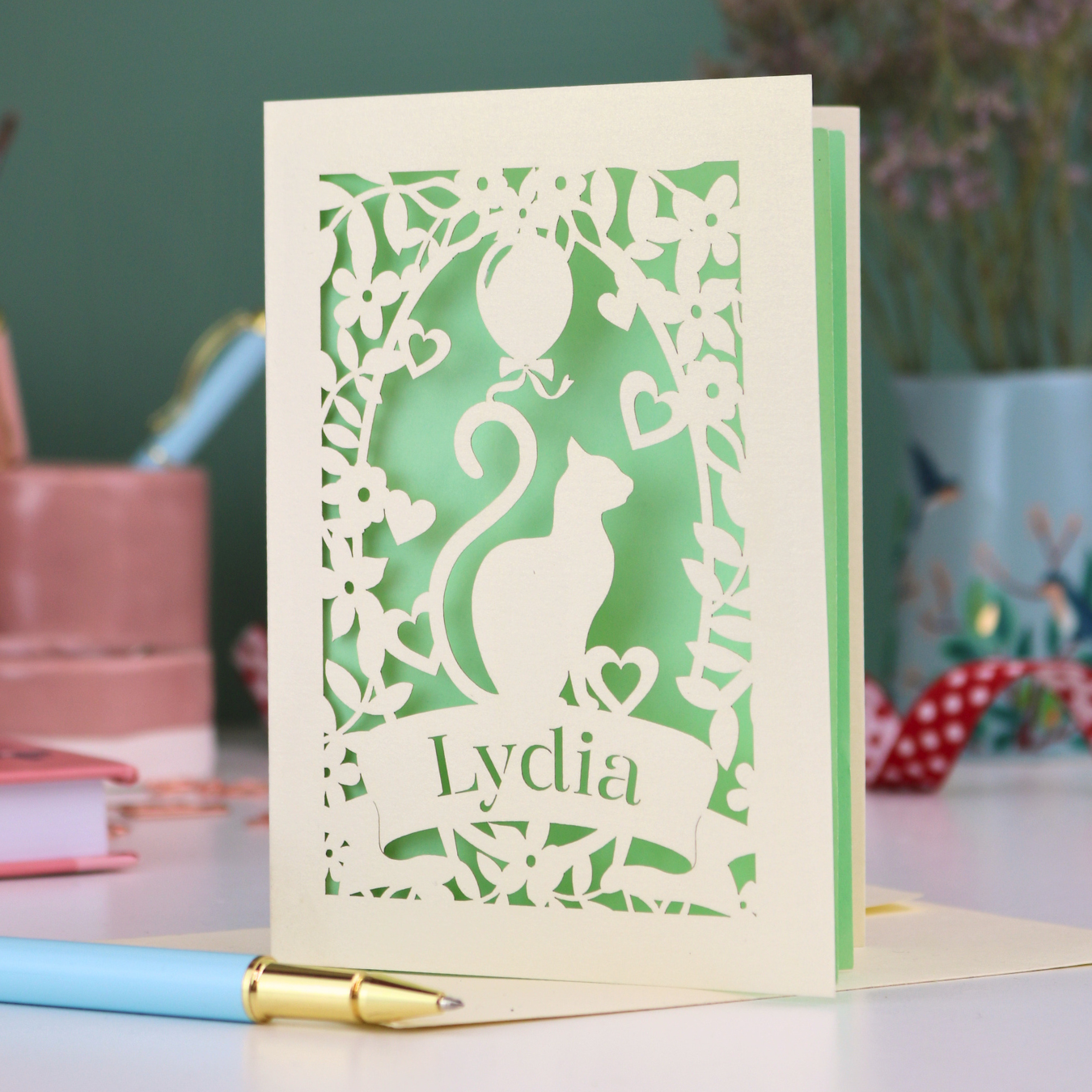A laser cut birthday card with cat design and personalised with a name, on a desk with stationery items. 
