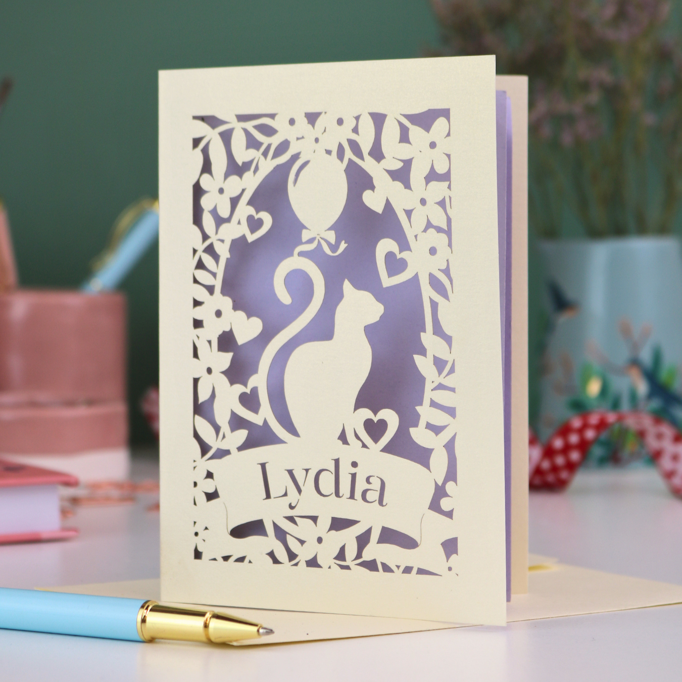 A laser cut birthday card with cat design and personalised with a name, on a desk with stationery items. 