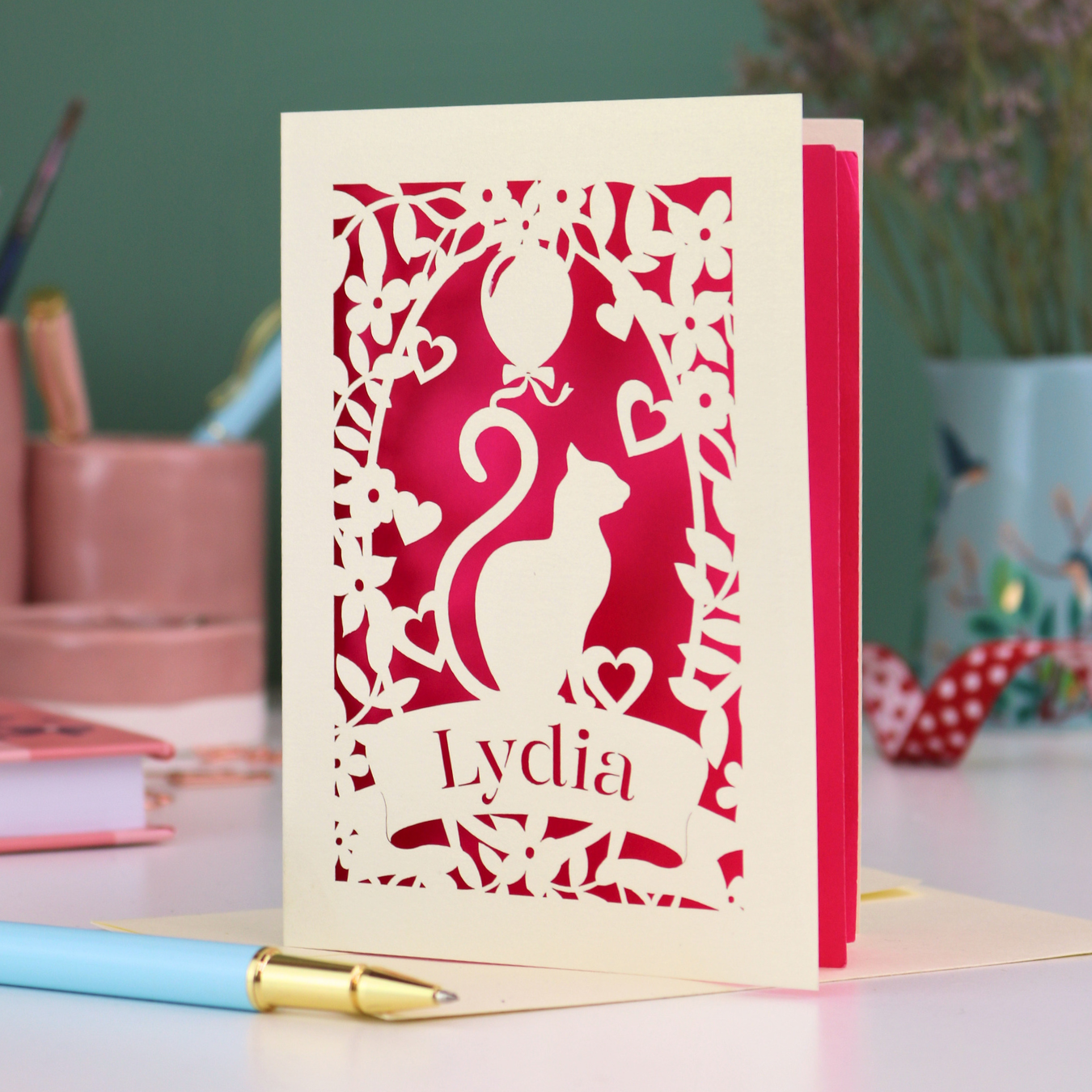 A laser cut birthday card with cat design and personalised with a name, on a desk with stationery items. 