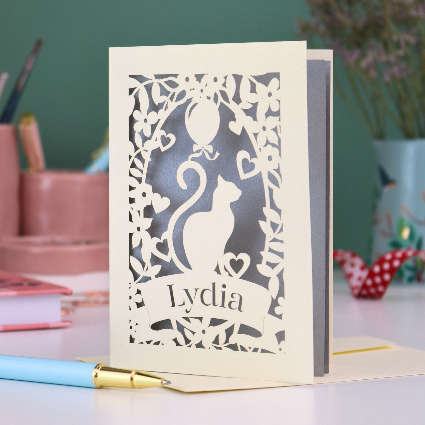 A laser cut birthday card with cat design and personalised with a name, on a desk with stationery items. 