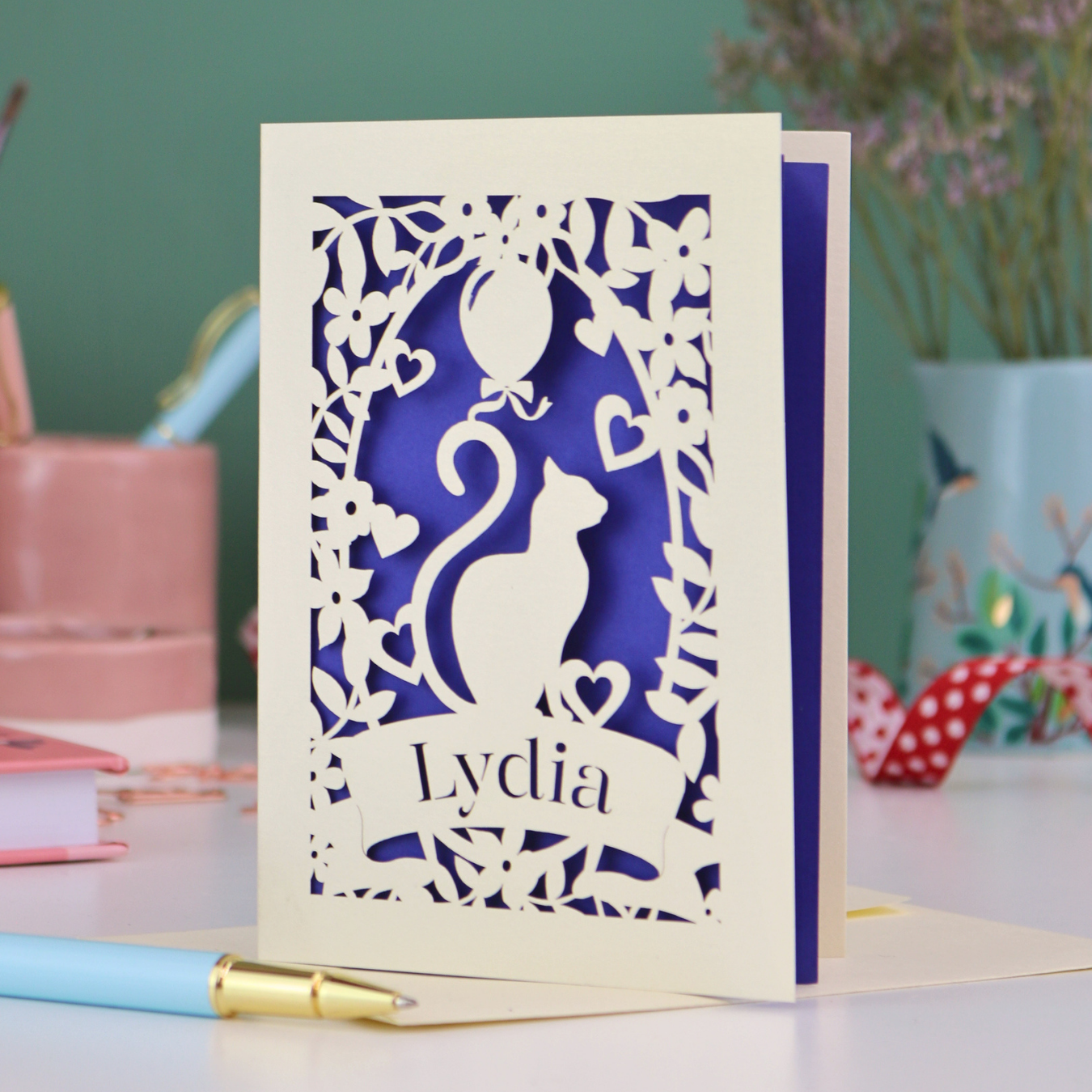 A laser cut birthday card with cat design and personalised with a name, on a desk with stationery items. 