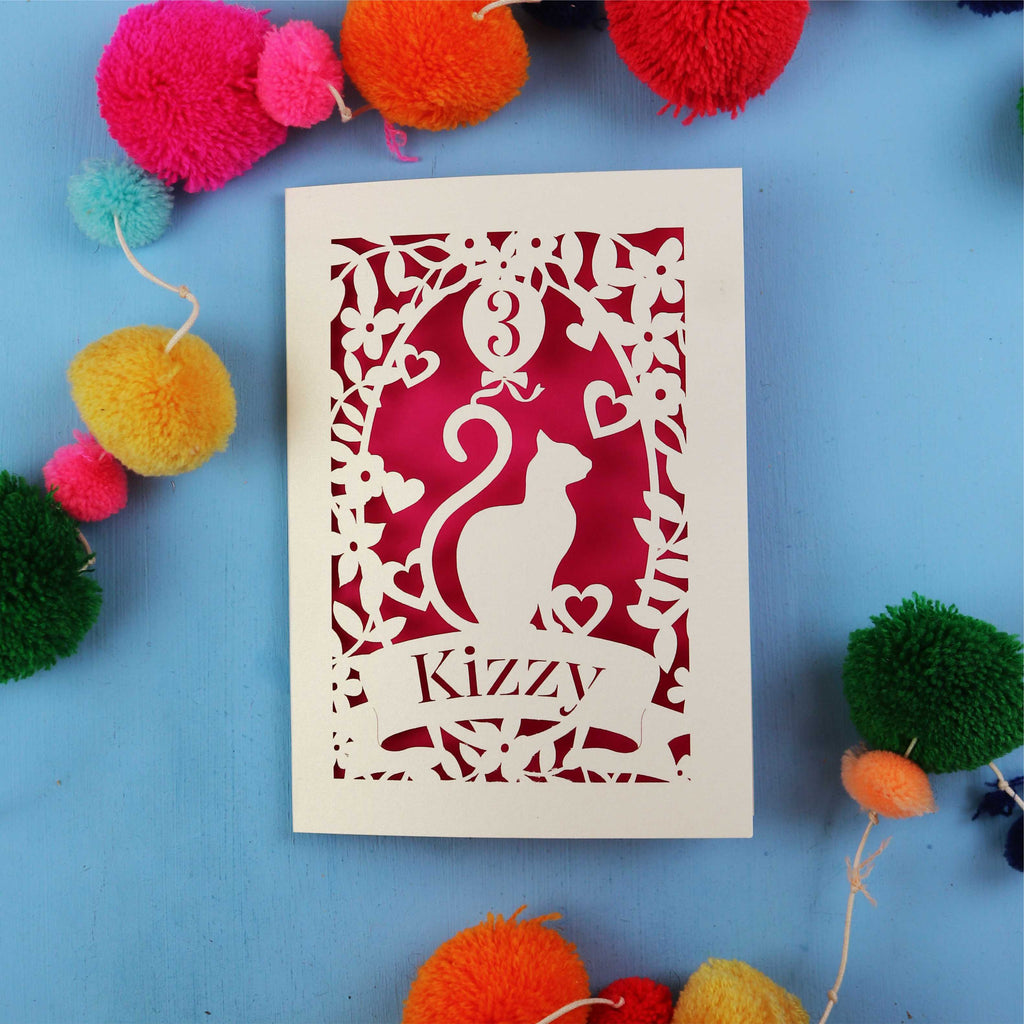 Decorative birthday card with cut-out design of a cat and text 'Kizzy' on a blue background with colourful pom-poms.
