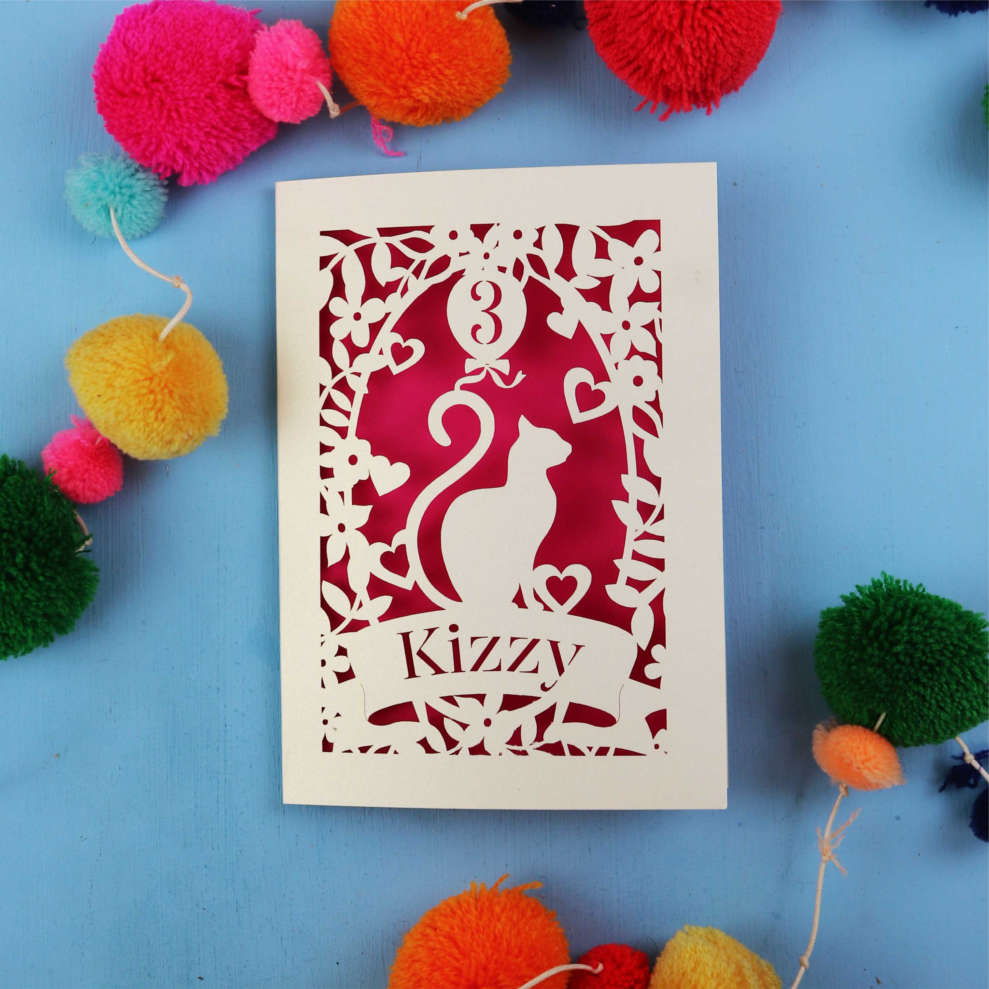 Decorative birthday card with cut-out design of a cat and text 'Kizzy' on a blue background with colourful pom-poms.