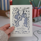Hand holding a birthday card with a cat design and 'Indie' text, set against a desk background.