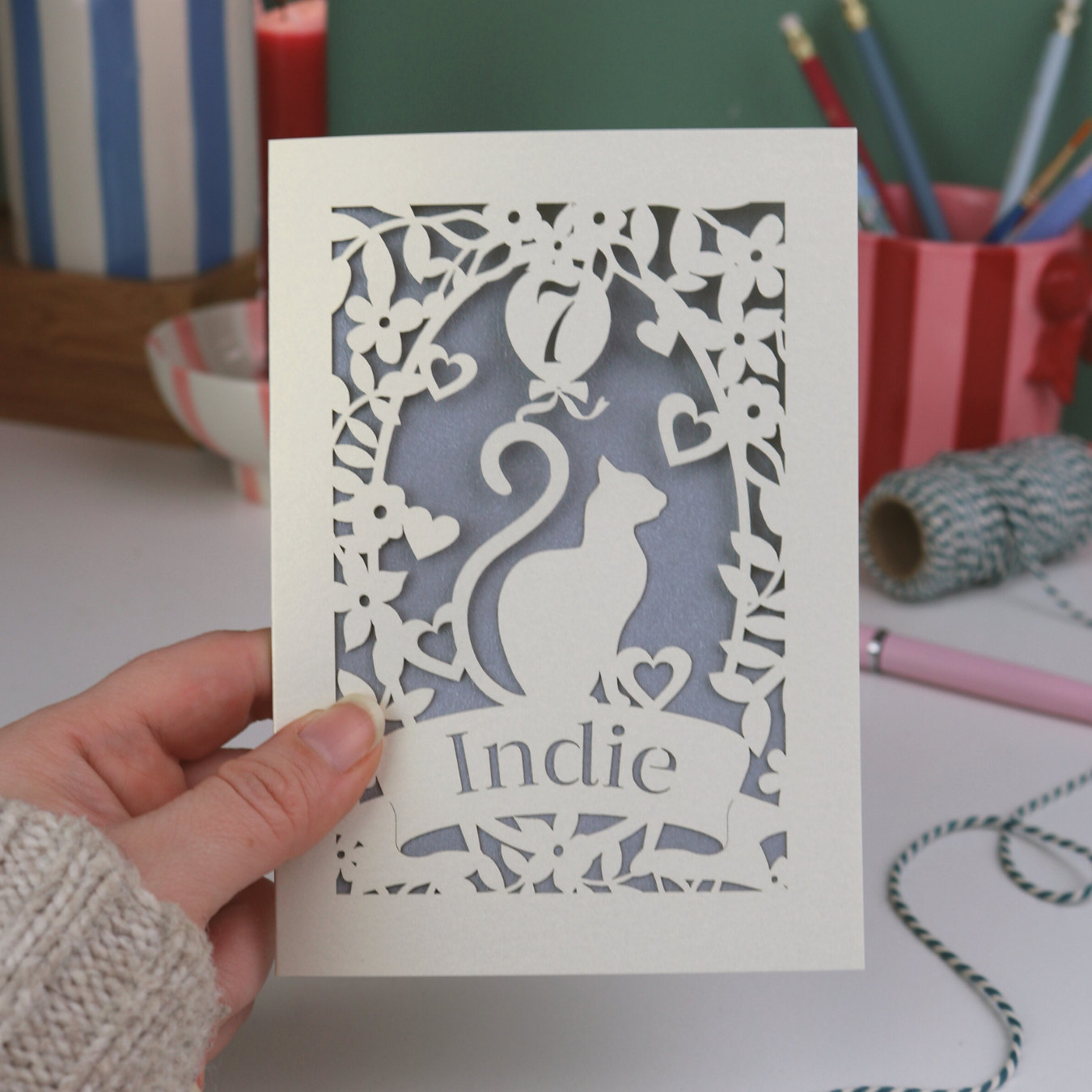 Hand holding a birthday card with a cat design and 'Indie' text, set against a desk background.