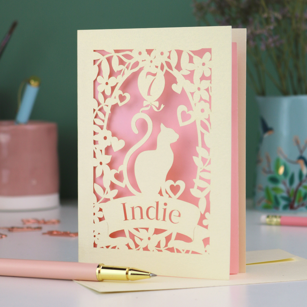 A cream laser cut birthday card with cat design and personalised with '7' in a balloon and the name of the recipient, 'Indie' in a banner