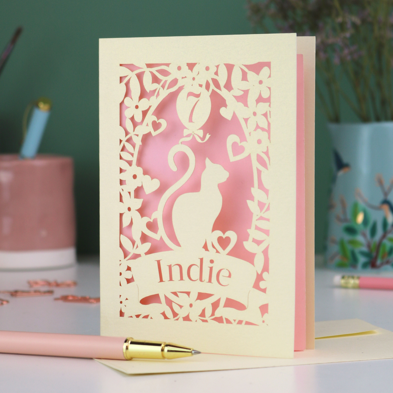 A cream laser cut birthday card with cat design and personalised with '7' in a balloon and the name of the recipient, 'Indie' in a banner