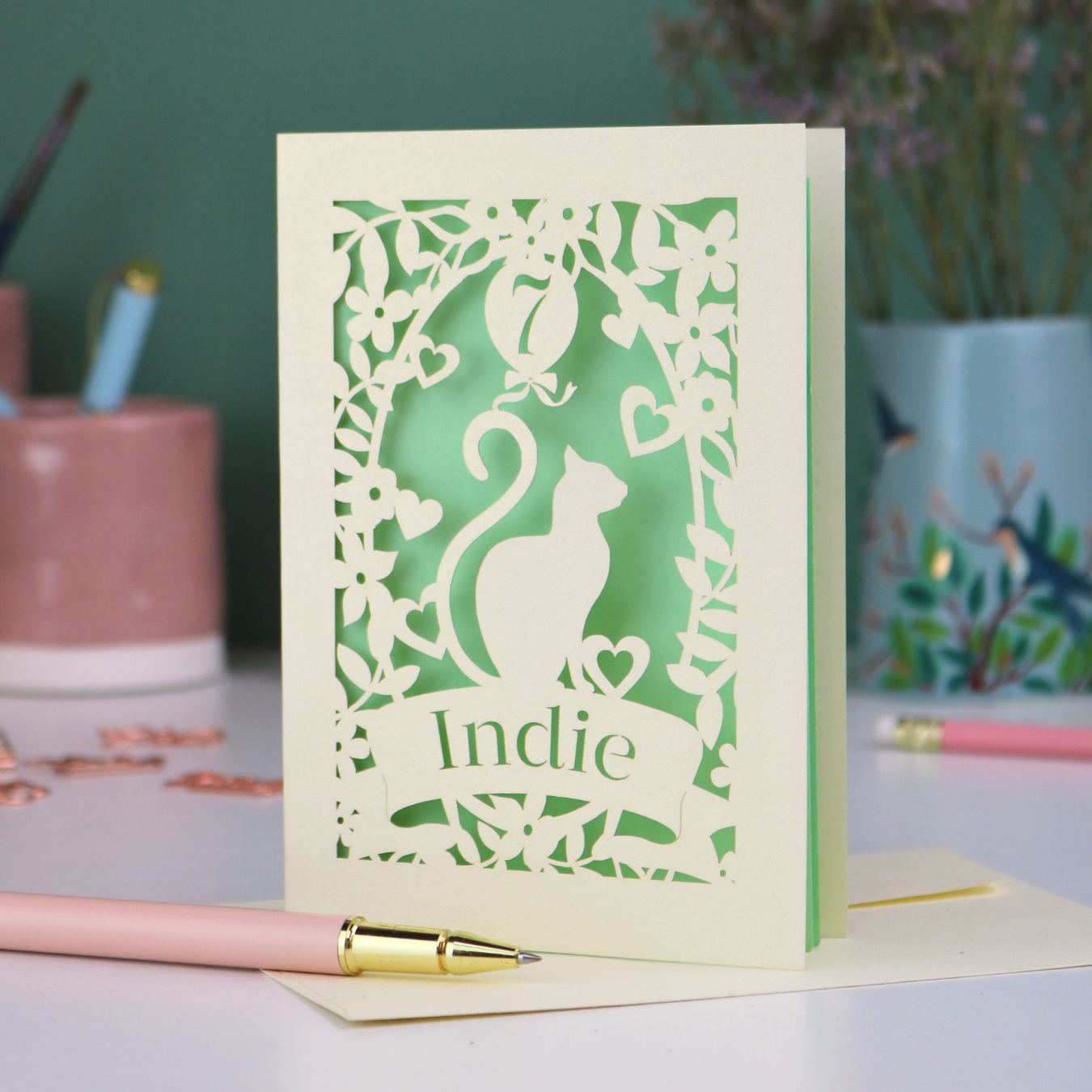 A cream laser cut birthday card with cat design and personalised with '7' in a balloon and the name of the recipient, 'Indie' in a banner