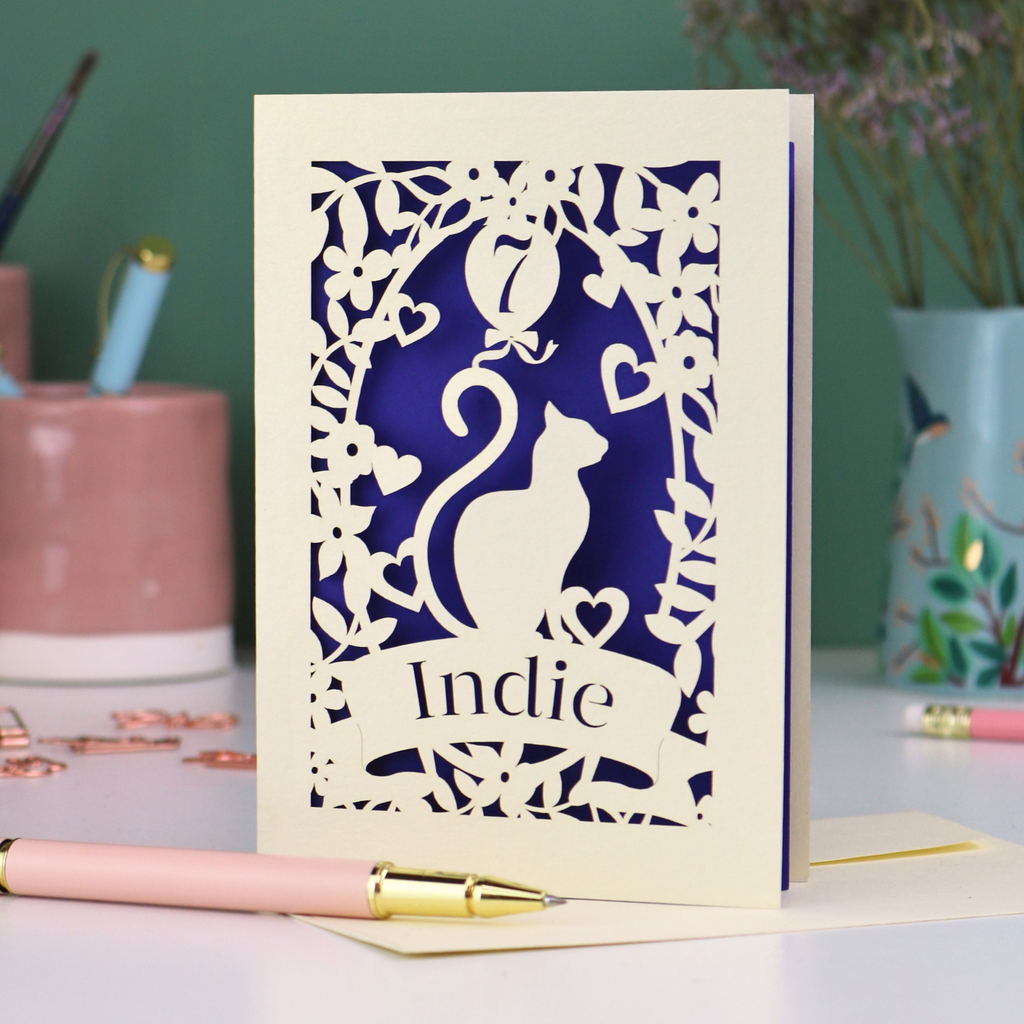 A cream laser cut birthday card with cat design and personalised with '7' in a balloon and the name of the recipient, 'Indie' in a banner