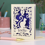 A cream laser cut birthday card with cat design and personalised with '7' in a balloon and the name of the recipient, 'Indie' in a banner