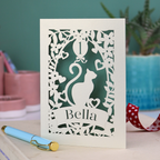 1st Birthday card with cat design and 'Bella' text on a table with a pen and plants in the background