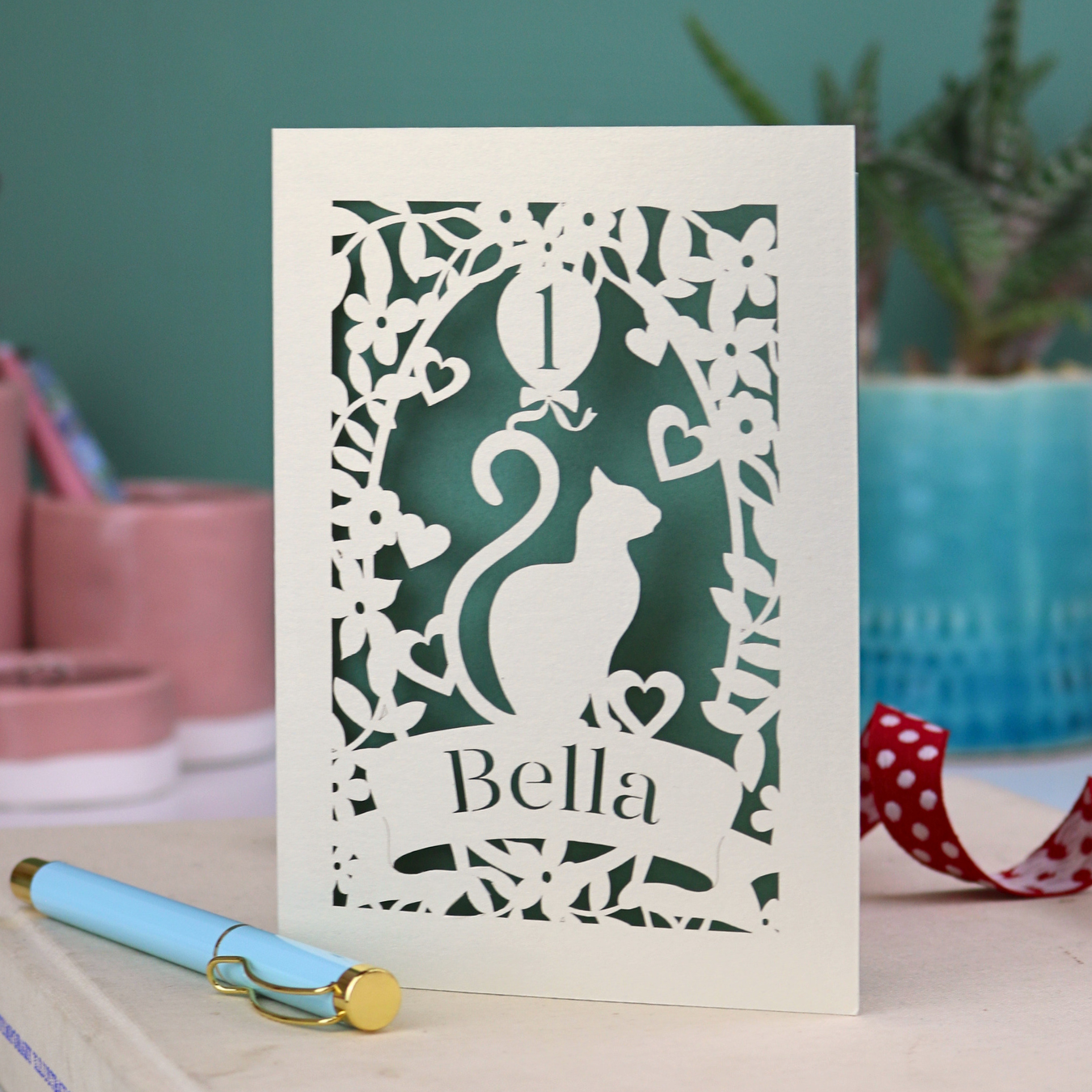 1st Birthday card with cat design and 'Bella' text on a table with a pen and plants in the background