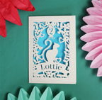 A cream laser cut birthday card with a cat and a balloon. The balloon has the number "2" cut out of it. There is a banner underneath the cat with a name cut out of it. The cut away design reveals a blue paper insert. 