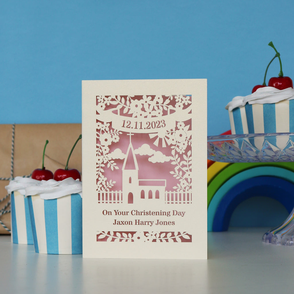 A cream card with a pink paper insert. This is a Christening card with the date and name personalised and a church in the centre.