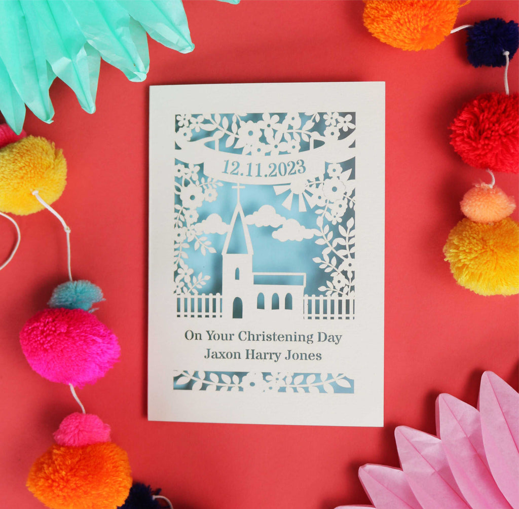 A laser cut card for a Christening. Card design is of a church surrounded by a border of flowers and leaves. There is a banner with a date at the top and under the church are the words "On your Christening Day" with a name underneath. Card is laser cut from cream card with a light blue paper insert behind the cut out card.