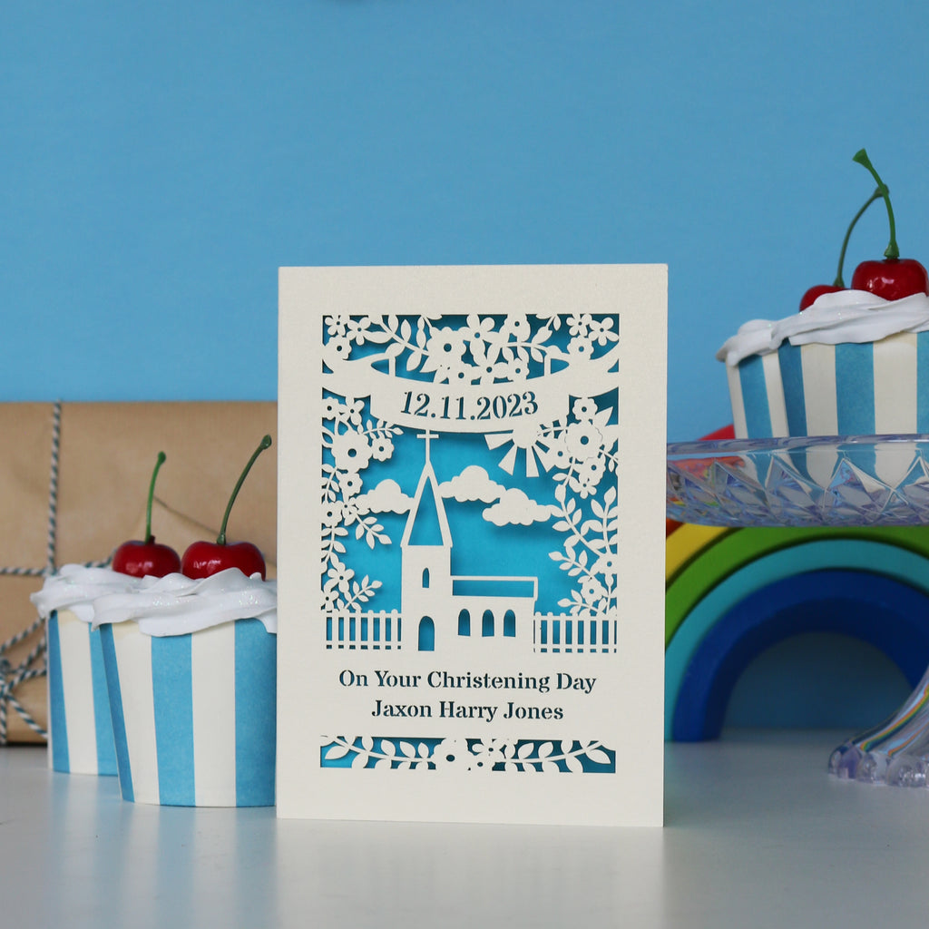 A cream and blue card for Christenings. Cut away card reveals blue paper behind and forms the shape of a church and flowers. Cut away text has the date at the top, "On Your Christening Day" and the name at the bottom.