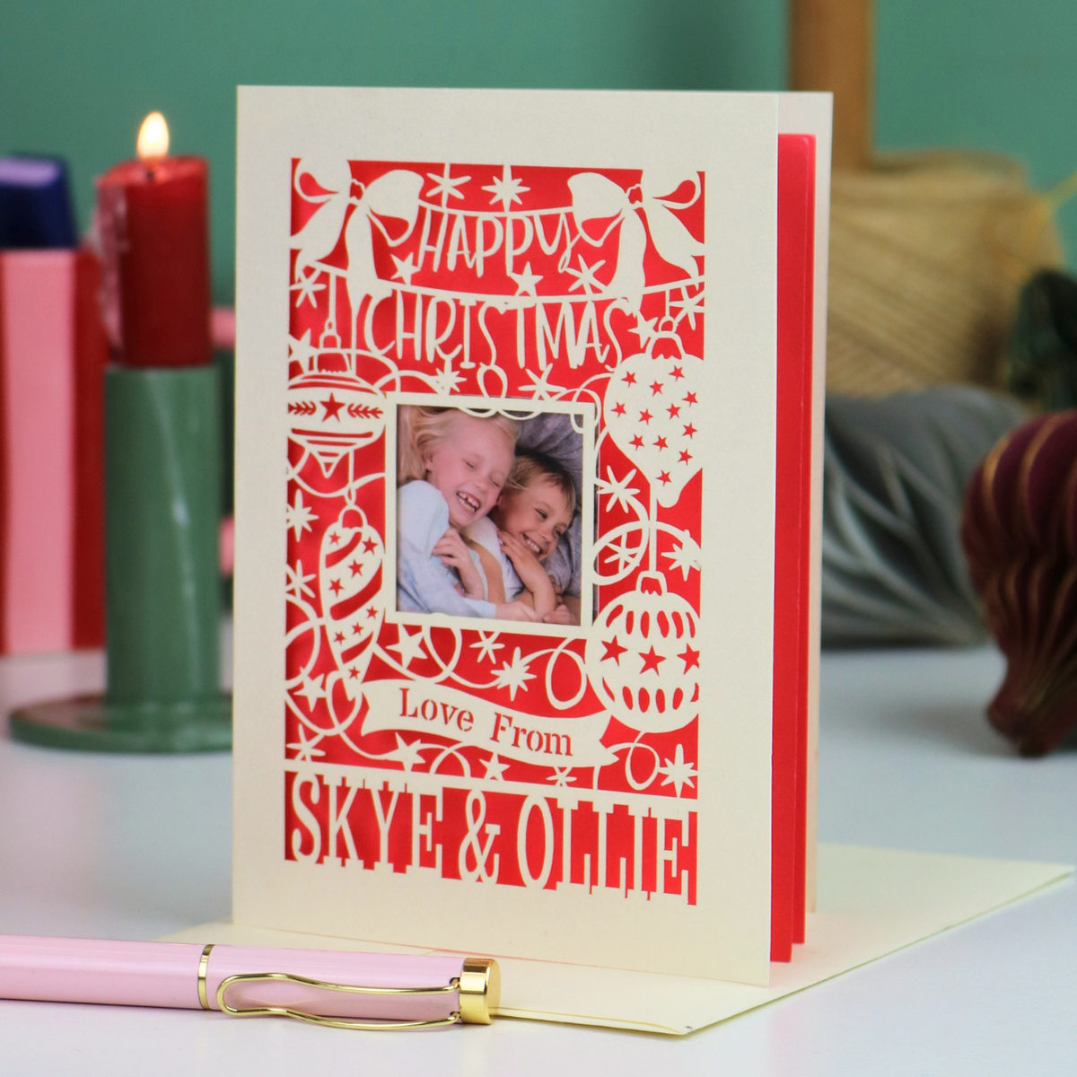 A personalized laser-cut cream-coloured Christmas card with coral backing and a photo of two children, displayed on a table with a pink pen in the foreground and holiday candles in the background.