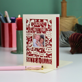 A personalized Christmas greeting card with a laser-cut red design on an off-white background, featuring a photo frame with two children and the text 'Happy Christmas Love From SKYE & OLLIE'. The card is displayed on a desk with a pink pen in the foreground and a blurred background with candles and stationery.