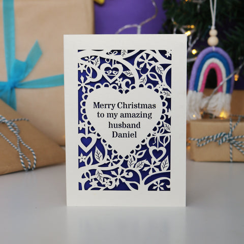 A personalised card for Christmas. Card has text inside a heart shape, with a border of hearts, flowers and snowflakes. Text reads "Merry Christmas to me amazing husband Daniel" - A5 / Cream / Infra Violet