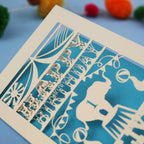 laser cut 1st Birthday card with cut-out design on a blue background with colourful pom-poms