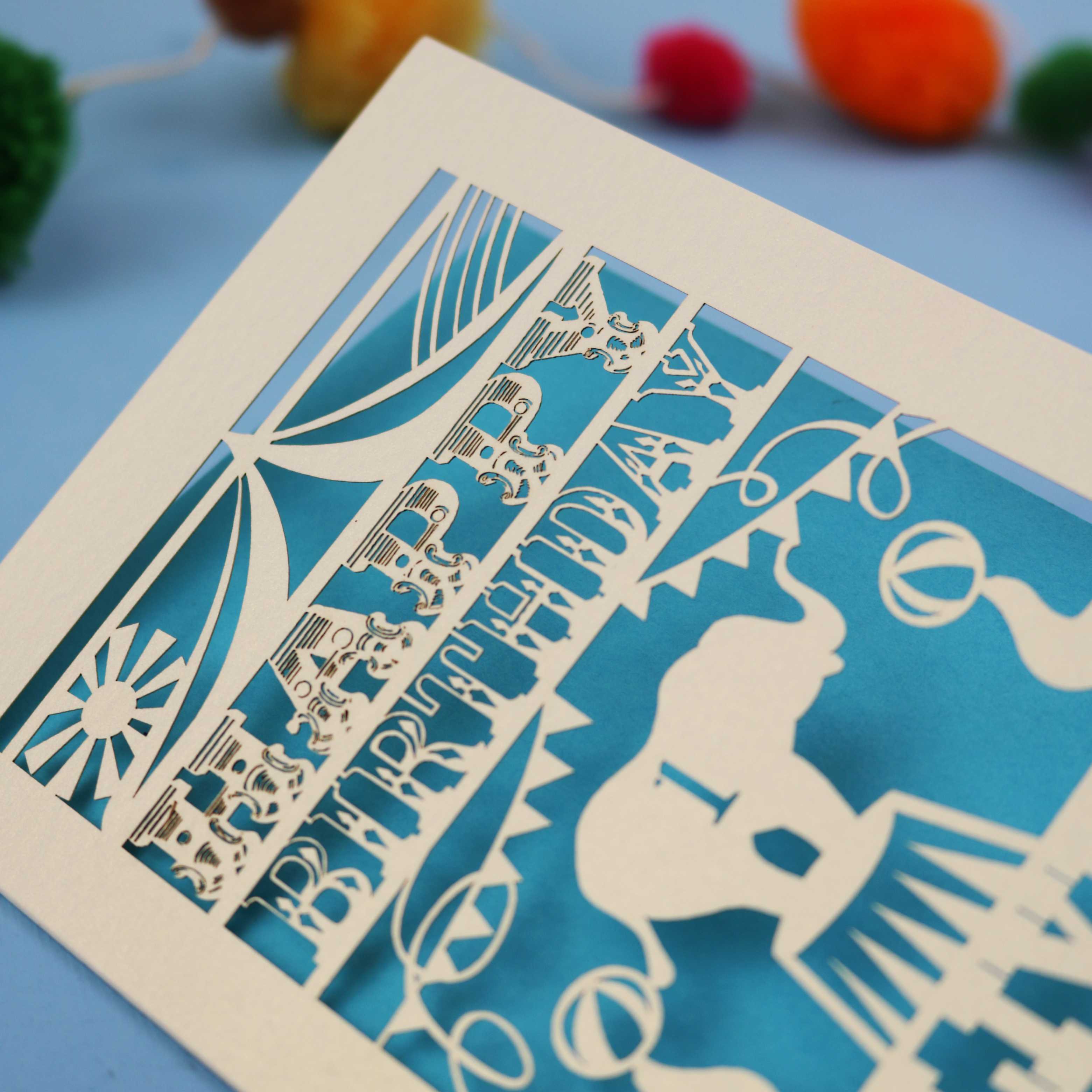 laser cut 1st Birthday card with cut-out design on a blue background with colourful pom-poms
