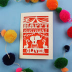 Paper cut First Birthday card with cut-out circus design on a blue background with colourful pom-poms