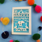 Paper cut First Birthday card with cut-out circus design on a blue background with colourful pom-poms