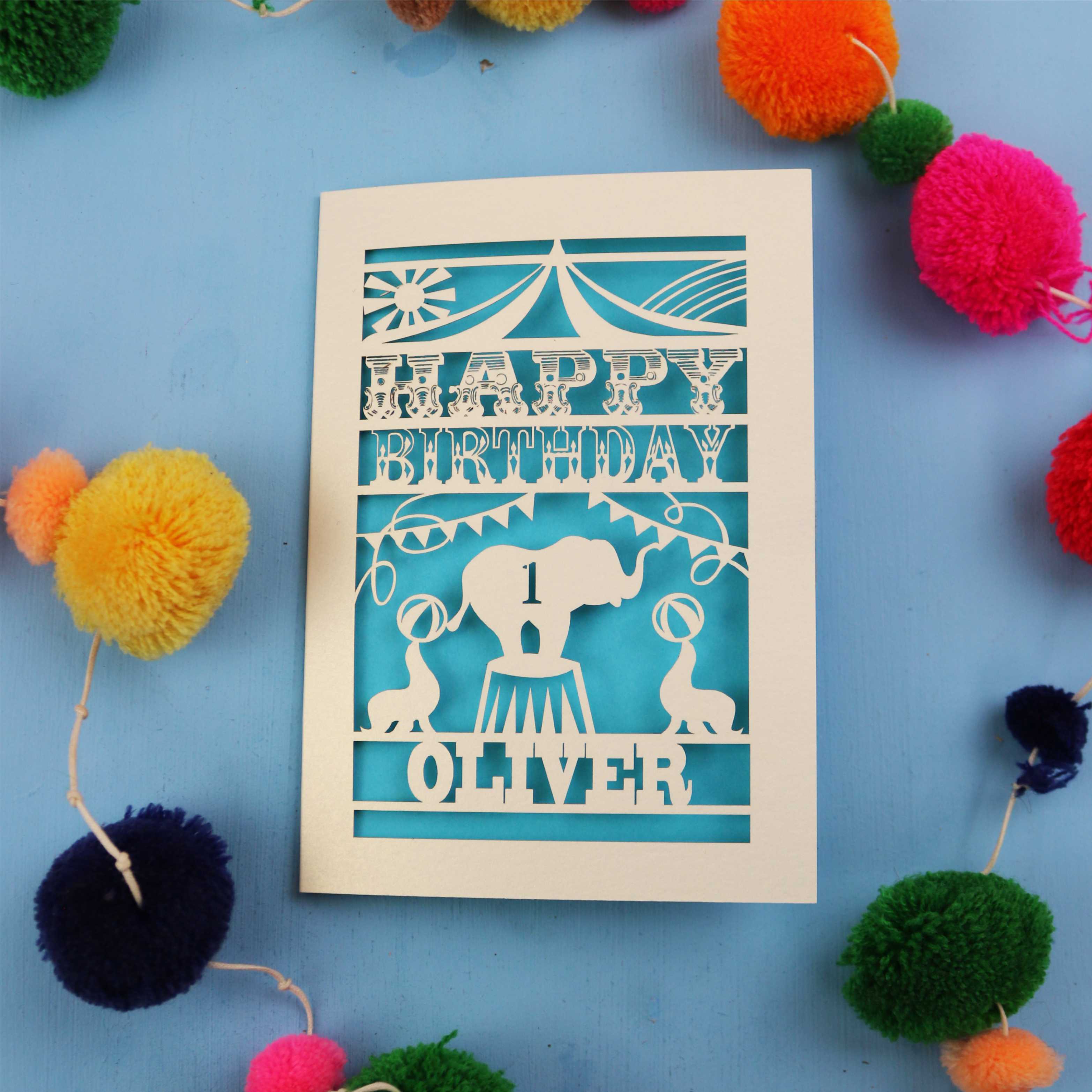 Paper cut First Birthday card with cut-out circus design on a blue background with colourful pom-poms