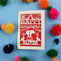 Paper cut First Birthday card with cut-out circus design on a blue background with colourful pom-poms