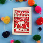 Paper cut First Birthday card with cut-out circus design on a blue background with colourful pom-poms
