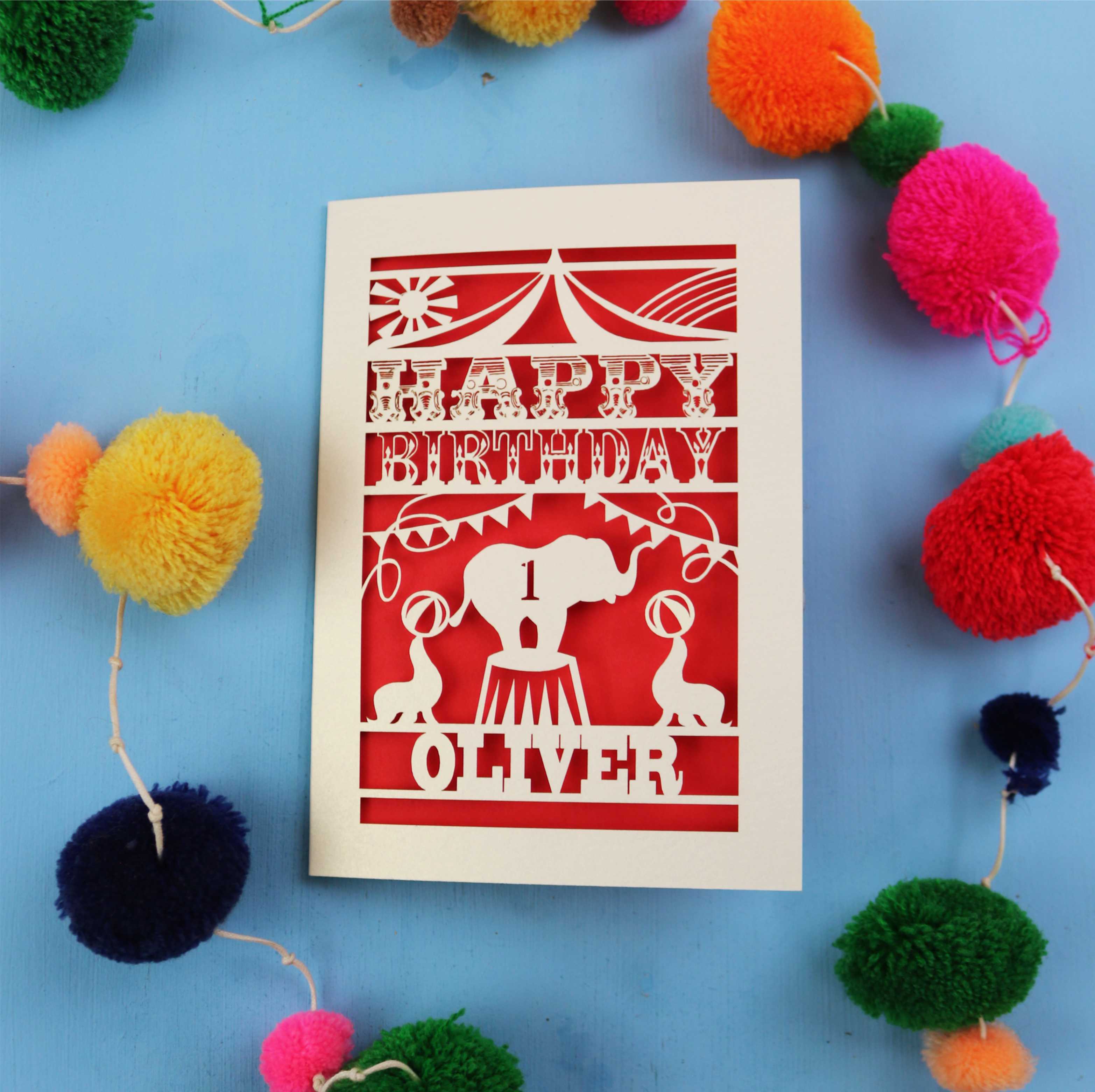 Paper cut First Birthday card with cut-out circus design on a blue background with colourful pom-poms