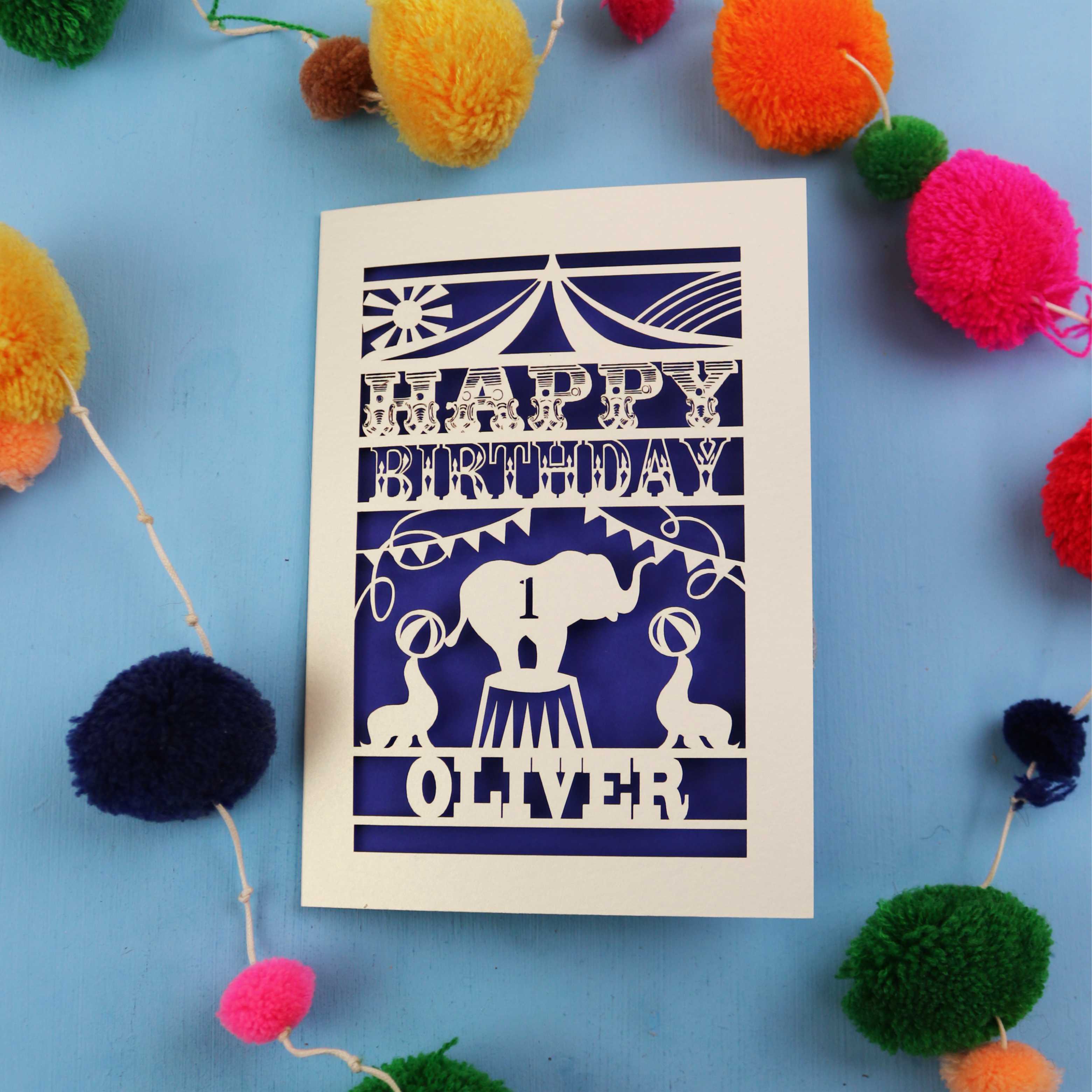 Paper cut First Birthday card with cut-out circus design on a blue background with colourful pom-poms