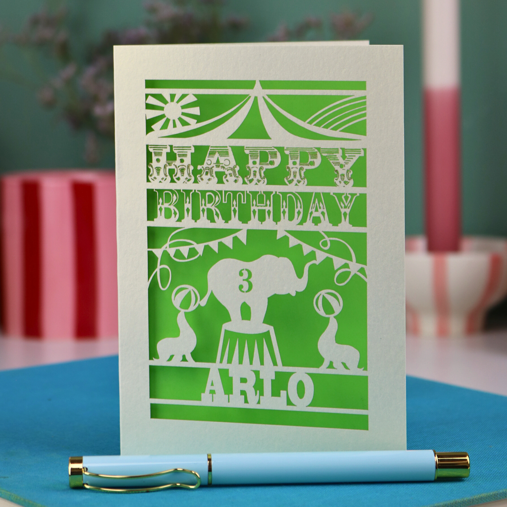 A laser cut birthday card with circus big top design, "Happy Birthday" and a name, with an age cut out.