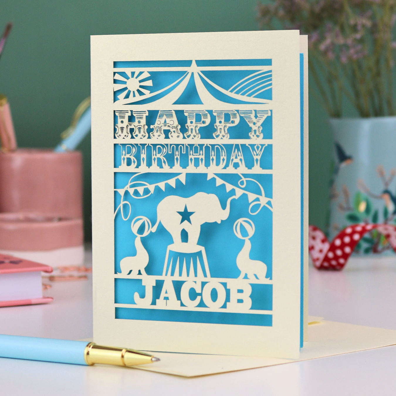 A laser cut personalised birthday card with circus elephant and sealions design and text "Happy Birthday Jacob", in cream with a bright blue paper insert, on white desk with stationery items