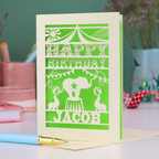 A laser cut personalised birthday card with circus design and text "Happy Birthday Jacob", in cream with a bright green paper insert, on white desk with stationery items