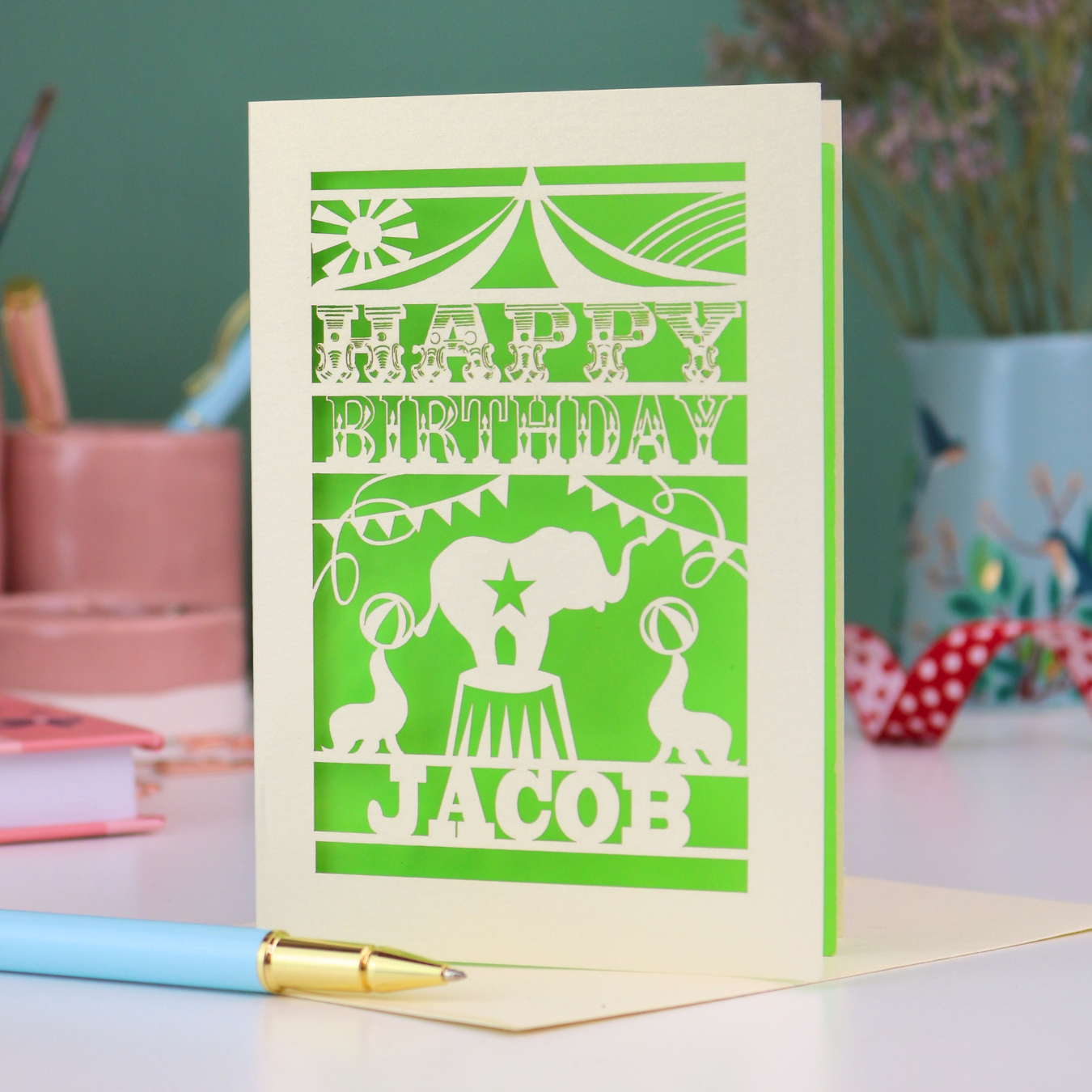 A laser cut personalised birthday card with circus design and text "Happy Birthday Jacob", in cream with a bright green paper insert, on white desk with stationery items