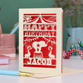 A laser cut personalised birthday card with circus design and text "Happy Birthday Jacob", in cream with a red paper insert, on white desk with stationery items
