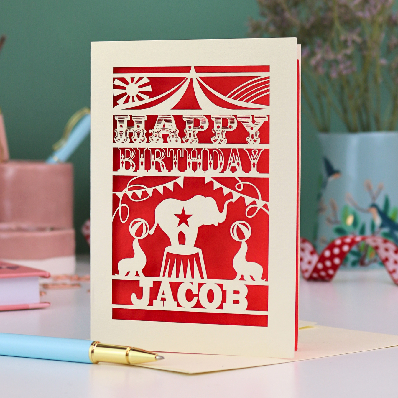 A laser cut personalised birthday card with circus design and text "Happy Birthday Jacob", in cream with a red paper insert, on white desk with stationery items